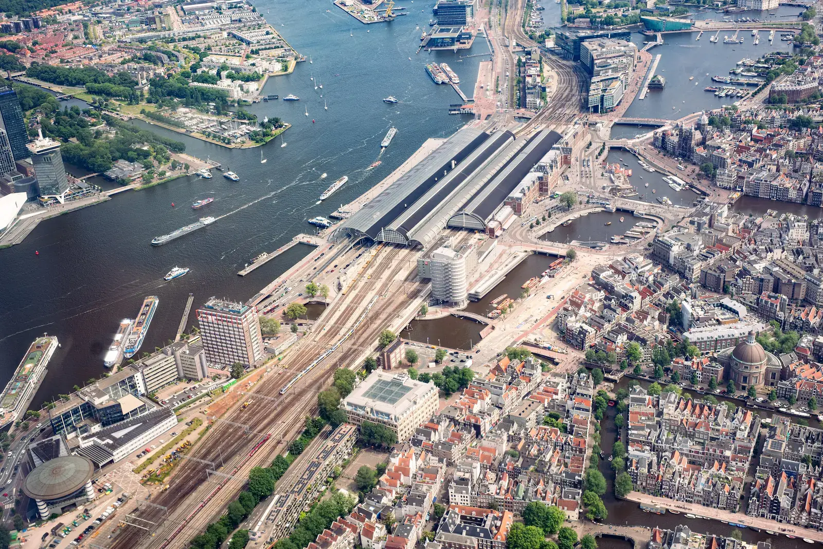 Luchtfoto van Amsterdam met het Centraal Station, het IJ en de dicht bebouwde binnenstad eromheen, waarin de groene plekken tussen de bouwblokken duidelijk te tellen zijn.