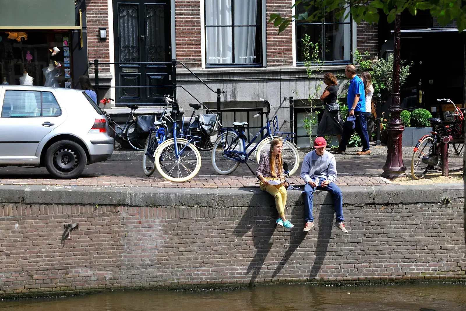 Historische bakstenen kademuur aan de Leliegracht in Amsterdam, met twee mensen op de rand, fietsen en een straatboom op de kade erboven.