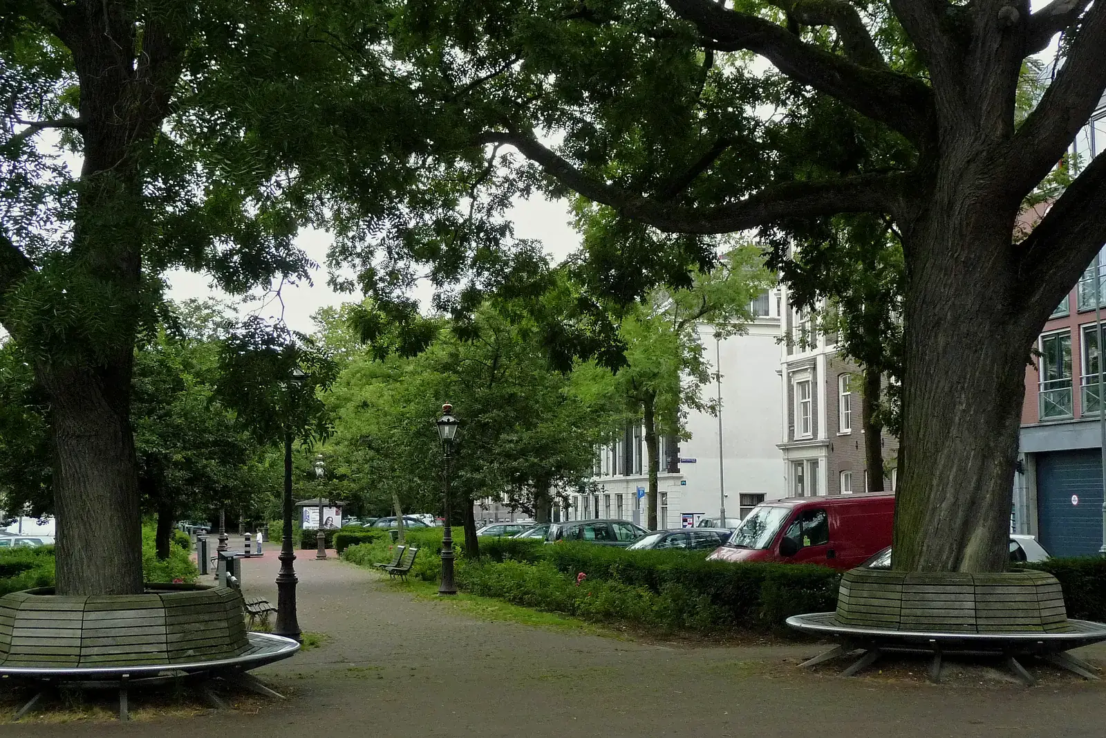 Volgroeide bomen op het Frederiksplein in Amsterdam werpen diepe schaduw over ronde banken, met geparkeerde auto's en gevels op de achtergrond.